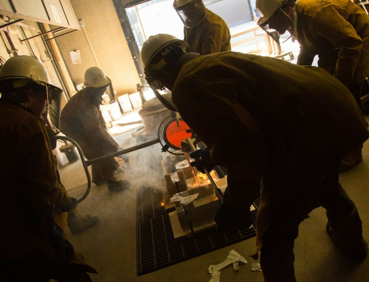 Darkened silhouettes of Penn State visual arts students crouched around a foundry crucible pouring red-hot molten metal.