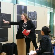 Student presenting work to reviewers, gesturing at floor plans.