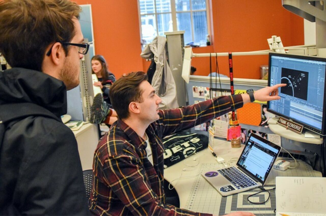 Two graphic design students looking at a design on an external monitor; one student is pointing a finger at a section of the screen.