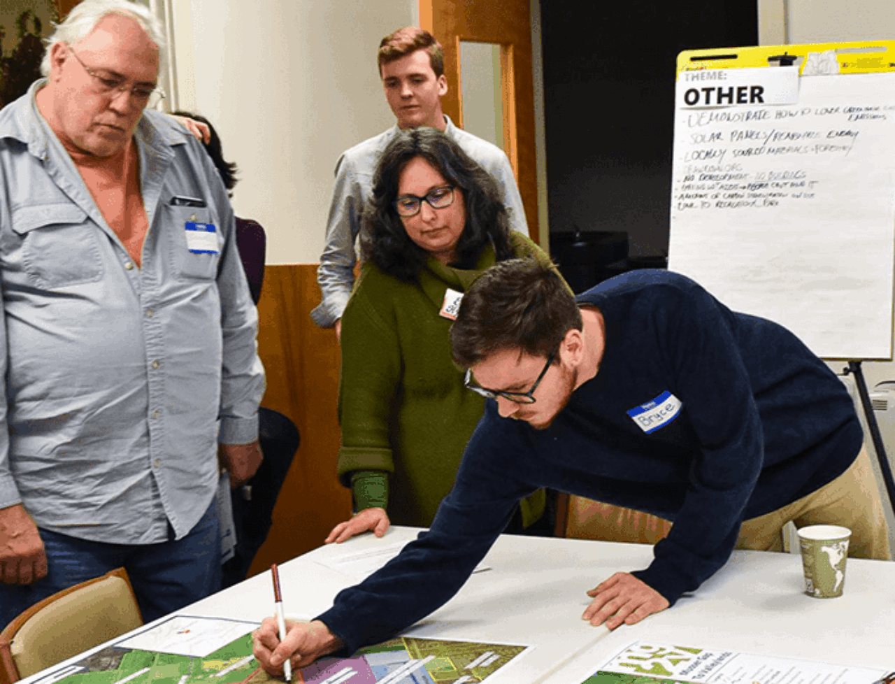 EplusD community meeting showcasing people watching a person drawing and making notes on a paper schematic on a table.