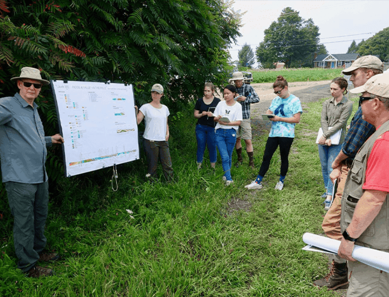 EplusD field work showcasing a group of researchers standing and listening intently to two researchers presenting information on a white board.