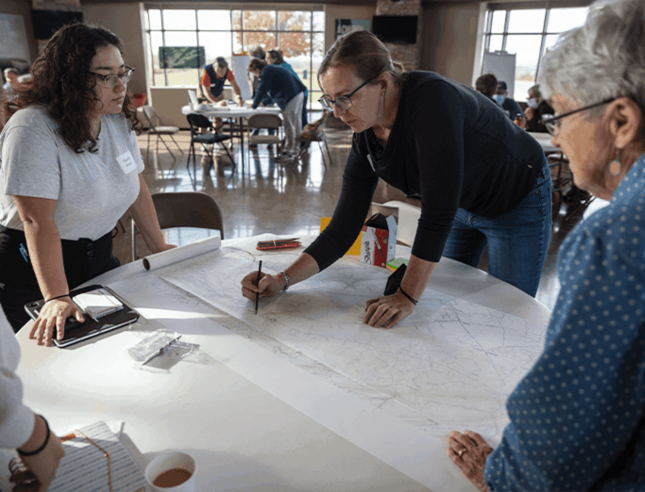 A community member leans over a map on the table and points something out to a student at left and another resident at right.