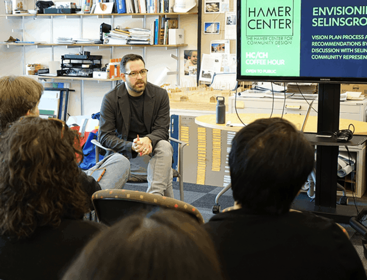 Stephen Mainzer sits facing community members during a community meeting with the Hamer Center logo behind him on a screen.