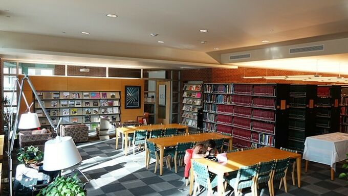 Inside view of the library with window light coming in from the left, students working at tables in the foreground, meeting space in the back, and stacks of books to the right.