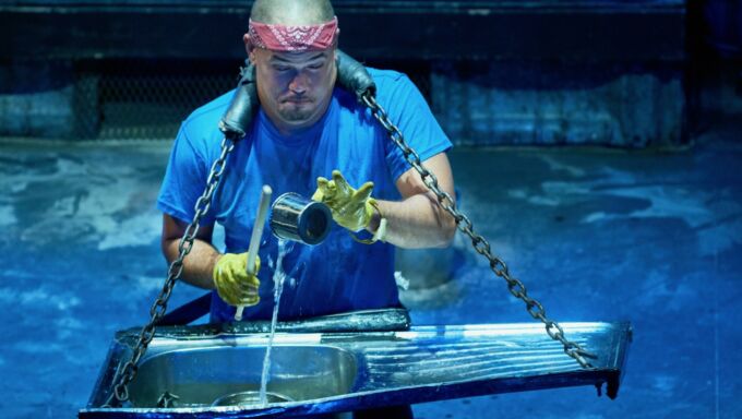 A bald man wears a stainless steel sink on a strap around his neck and he pours water into the bowl.