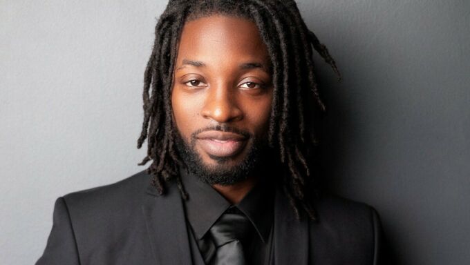 A Black man wearing short dreadlocks looks pleasantly chill at the camera.