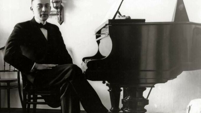 An historic photograph shows a man wearing a suit and bowtie and sitting at the keys of a piano.