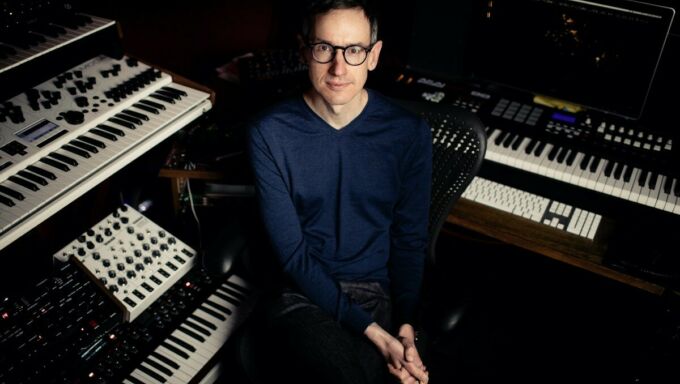 A man wears round black-rimmed glasses and sits surrounded by high-tech keyboards and synthesizers.