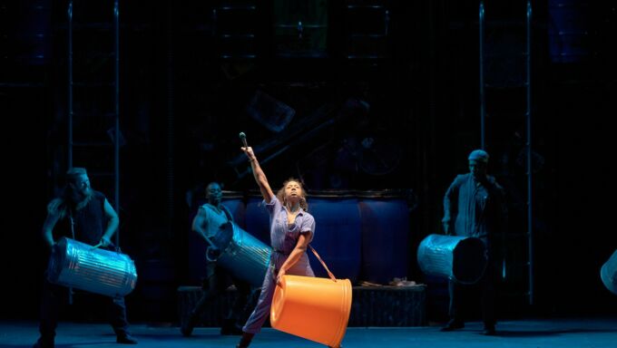 A woman of color standing center stage wears a plastic drum on a strap around her neck and shoulders as she points her arms upward.