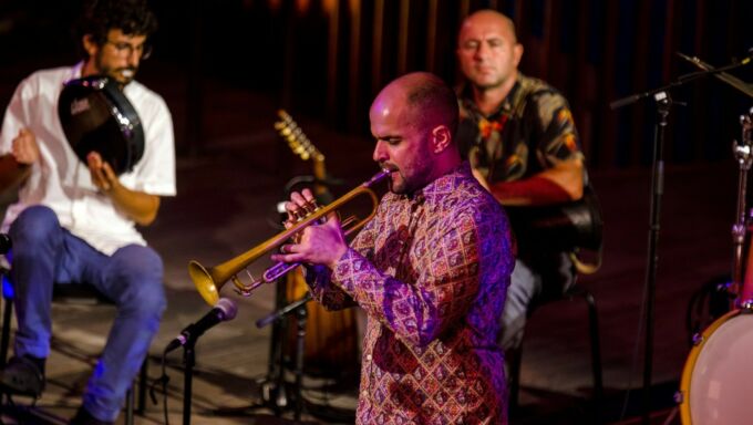 A man plays a trumpet while two percussion musicians in the background accompany.