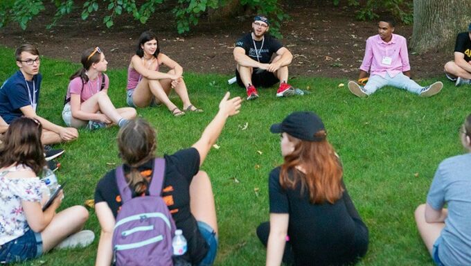 Summer camp participants sitting in a circle on a patch of grass talking.