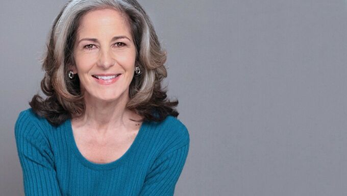 Photo of a woman with greying hair wearing a blue shirt and smiling in front of a grey background