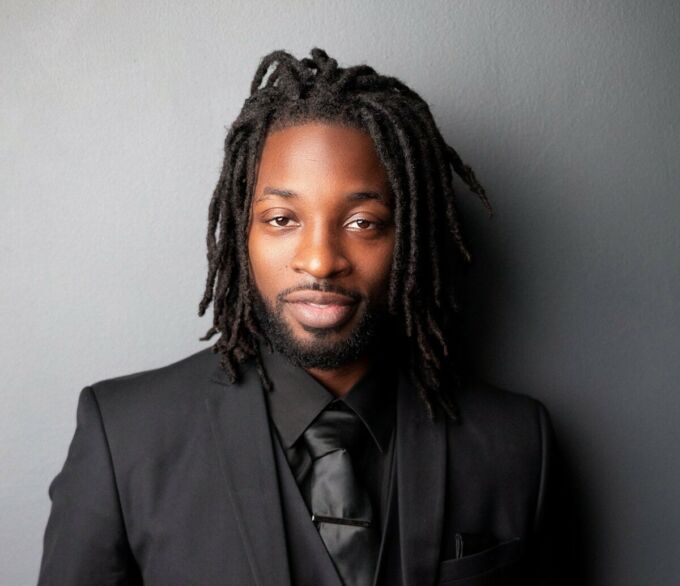 A Black man wearing short dreadlocks looks pleasantly chill at the camera.