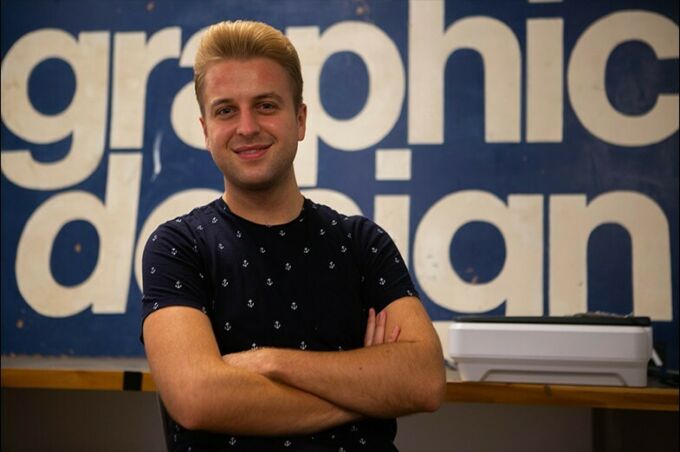 Portrait of Jeff Buterbaugh standing with his arms folded in front of the graphic design sign in the senior studio space.