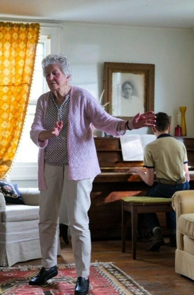 An elderly woman dancing with a man behind her at a piano