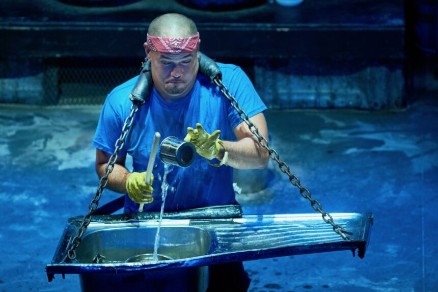 A bald man wears a stainless steel sink on a strap around his neck and he pours water into the bowl.