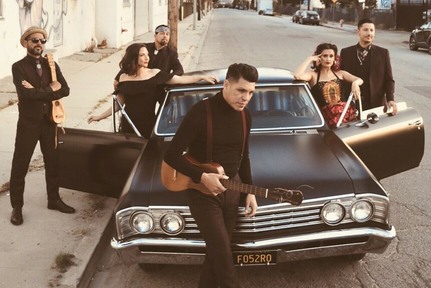 A group of men and women dressed monochromatically stand glamourously around a souped up hot rod.