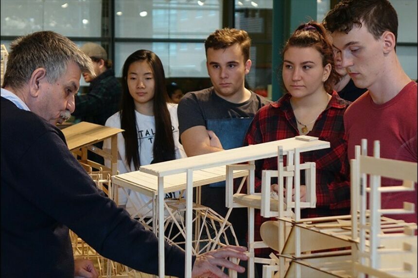 A side profile of Loukas Kalisperis pointing at a model on a desk in front of him while students watch.