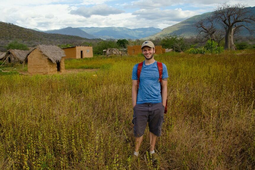 Man in blue shirt and grey shorts standing in front of grass-thatched huts with the mountains of Tanzania towering in the background.