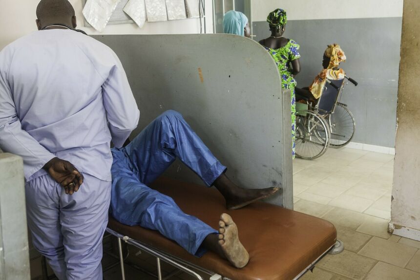 Doctor examining patient lying on a gurney behind a partition. In the background, two women are standing adjacent to a wheelchair-bound woman.