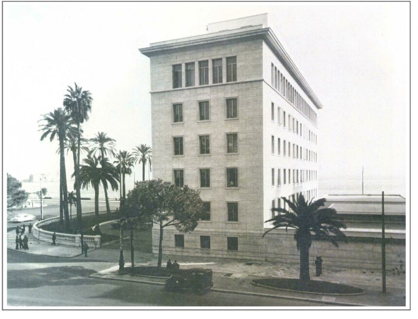 Old black and white photo of a six-story building by the waterfront with palm trees dotting its property and pedestrians walking past.